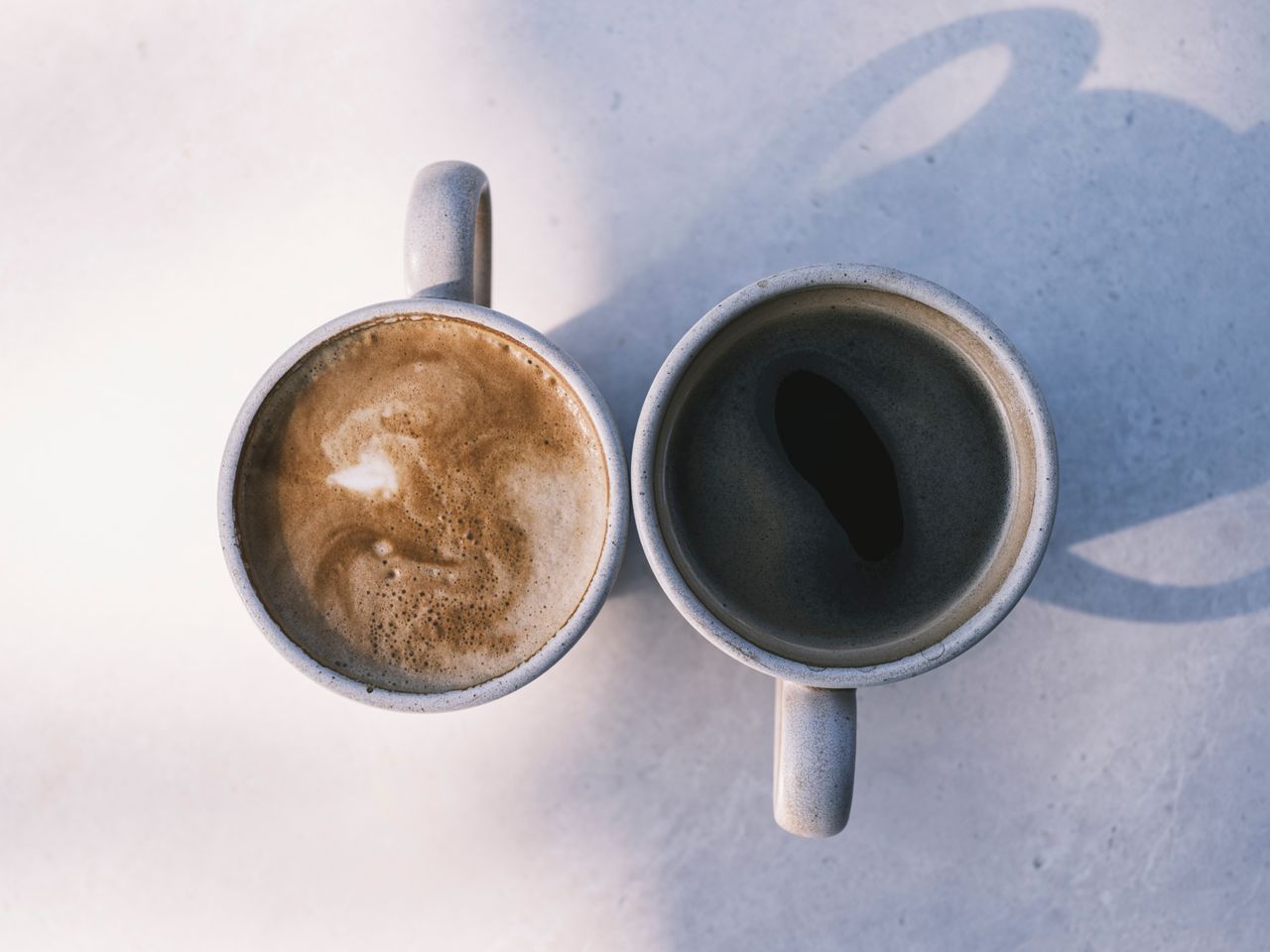 Services Aerial view of two coffee cups casting shadows on a sunny table, showcasing latte and black coffee.